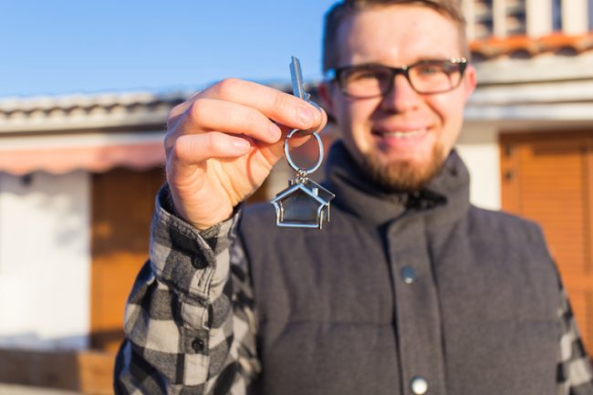 Dwelling, buying home, real estate and ownership concept – handsome man showing his key to new home man holding key to new home